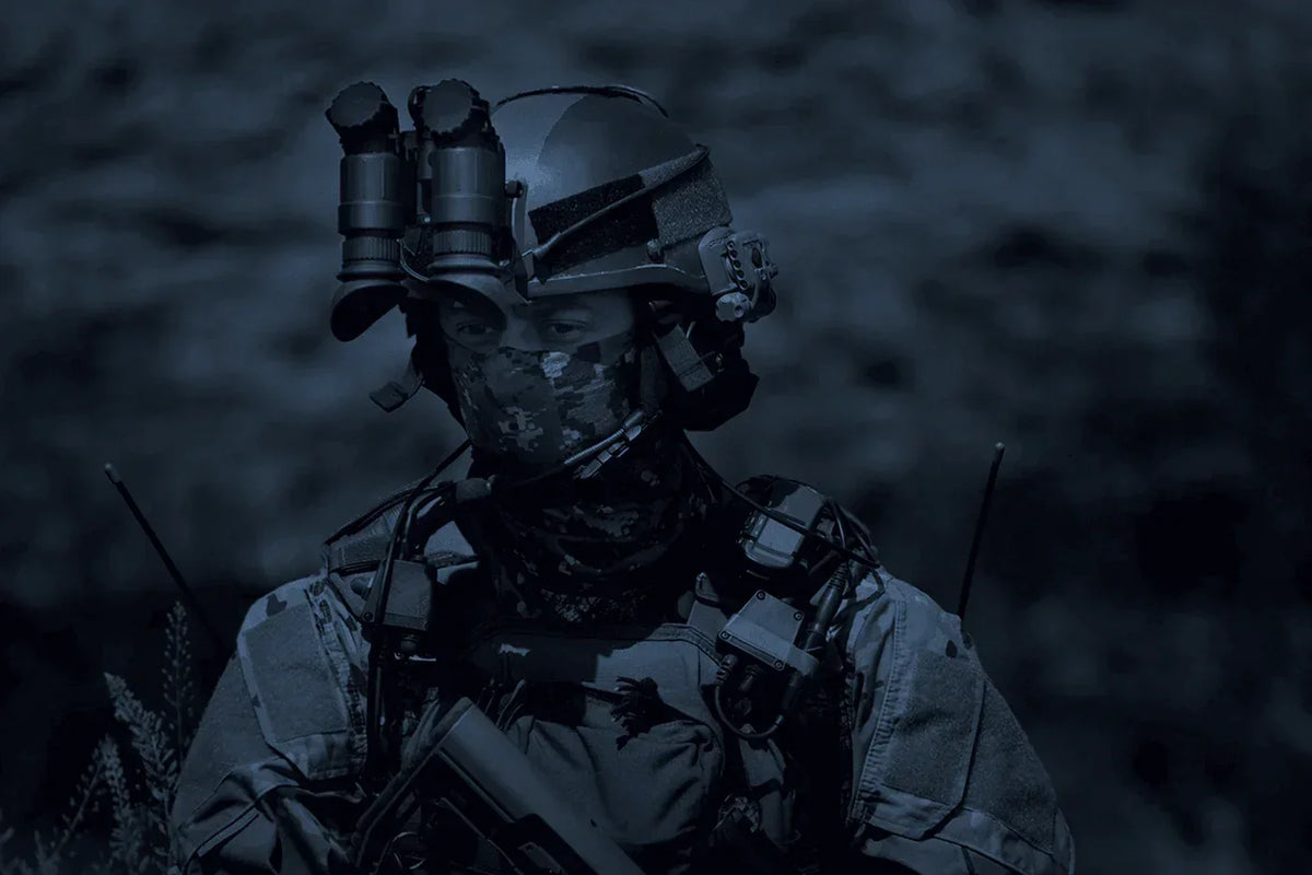 Soldier wearing helmet-mounted night vision binoculars, tactical gear, and camouflage face covering